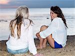 Two young women friends sitting chatting on beach at sunset, Williamstown, Melbourne, Australia