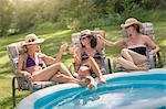 Three mature women relaxing in sun loungers and drinking wine, next to paddling pool