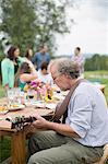 Mature man playing guitar while friends and family talk together after meal, outdoors