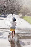 Rear view of barefoot girl carrying umbrella walking through street puddle