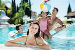 Portrait of smiling woman in swimming pool, family playing with beach ball in background