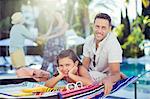 Portrait of smiling father and daughter relaxing by swimming pool, mother and daughter in background