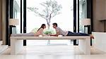 Couple lying on table face to face and holding hands, tree seen through patio door in background