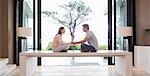 Couple sitting on table face to face and holding hands, tree seen through patio door in background