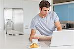 Man with newspaper using laptop at kitchen counter, coffee cup and croissant in foreground