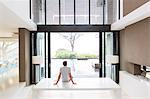 Rear view of man sitting on table and looking at swimming pool through open patio door