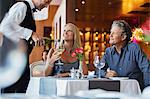 Mature couple sitting at restaurant table, waiter pouring white wine into woman's glass