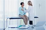 Female doctor examining her patient with stethoscope in office, desk with laptop in foreground