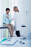 Female doctor examining her patient in office, laptop, otoscope, file and glasses on desk in foreground