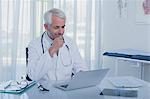 Smiling mature doctor sitting at desk with laptop in office