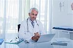 Portrait of smiling mature doctor sitting at desk with laptop in office