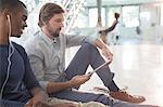 Two businessmen using digital tablet and earphones sitting on floor in modern office