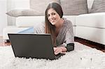 Portrait of smiling young woman with laptop lying on carpet at home