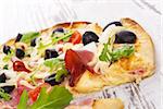 Delicious colorful pizza with tomatoes, melted cheese, black olives, onions, ham and fresh herbs on white wooden background. Culinary mediterranean pizza eating.