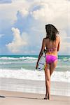 Rear view of a beautiful sexy young woman surfer girl in bikini with surfboard standing in the surf on a beach