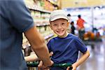 positive smiling boy sitting in the shopping cart while grocery shopping with his father together at supermarket