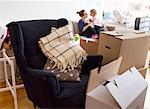 Boxes and armchair in apartment, mother with daughter in background