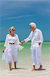 Happy senior man and woman couple walking, laughing and holding hands on a deserted tropical beach with bright clear blue sky