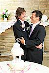 Two handsome gay men in tuxedos embrace with love at their wedding reception.  Wedding cake and rose petals in foreground.