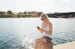 Young woman sitting at riverside texting on smartphone, Danube Island, Vienna, Austria