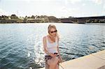 Young woman sitting at riverside reading texts on smartphone, Danube Island, Vienna, Austria