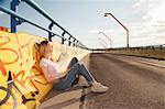 Young woman sitting leaning against graffiti wall on bridge in sunlight