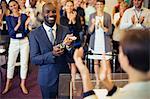 Portrait of young man holding trophy, standing in conference room, smiling to applauding audience