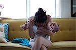 Mother holding and kissing baby's belly, sitting on sofa by window