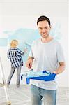 Portrait of man holding paint roller and tray with woman painting wall in background