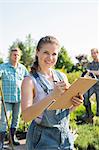 Portrait of beautiful supervisor holding clipboard with gardeners standing in background at plant nursery