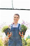 Portrait of happy female gardener holding potted plants at greenhouse