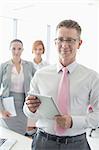 Portrait of happy businessman holding digital tablet with female colleagues in background at office