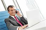 Smiling mature businessman talking on cell phone while using laptop in office