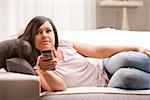 young woman watching tv on her sofa in her living room changing programme using a gray remote control