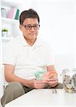 Portrait of mature Asian man counting on his savings, sitting on sofa at home, senior retiree indoors living lifestyle.