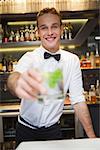 Happy bartender offering cocktail to camera in a bar