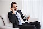 Businessman making a call on his couch reading newspaper at home in the living room