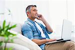 Cheerful man sitting on couch phoning and using laptop at home in the living room