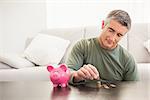 Smiling man putting some coins into a piggy bank at home in the living room