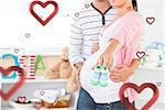 Close up of a bright pregnant woman holding baby shoes while husband touching her belly in the room  against hearts