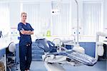 Dentist in blue scrubs standing with arms crossed beside chair at the dental clinic