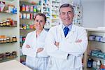 Smiling pharmacist and his trainee with arms crossed in the pharmacy