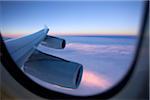 An aircraft wing and engines as seen from the window of a jet while flying above the clouds at sunset.