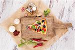 Culinary pizza cooking. Pizza piece on wooden kitchen board, with various ingredients and toppings. Fresh herbs, olive oil, garlic, edible mushrooms, dried tomatoes and chili pepper. Pizza eating.