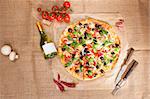 Country style pizza eating. Delicious round pizza with colorful various topping, prosciutto, ham, black olives and fresh herbs with wine bottle and ingredients on brown background, top view.