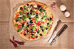 Round colorful pizza with ham, cheese, black olives, onion and various herbs on brown background, top view. Culinary pizza eating, rustic country style.