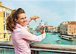 Portrait of happy young woman framing with hands while standing on bridge with grand canal view in venice, italy