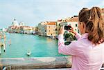 Young woman standing on bridge with grand canal view in venice, italy and taking photo. rear view