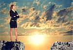 Businesswoman standing on the edge of rock gap, with her arms crossed on her breast as if uncertain. Sunrise sky as backdrop