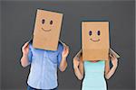 Couple wearing emoticon face boxes on their heads against grey